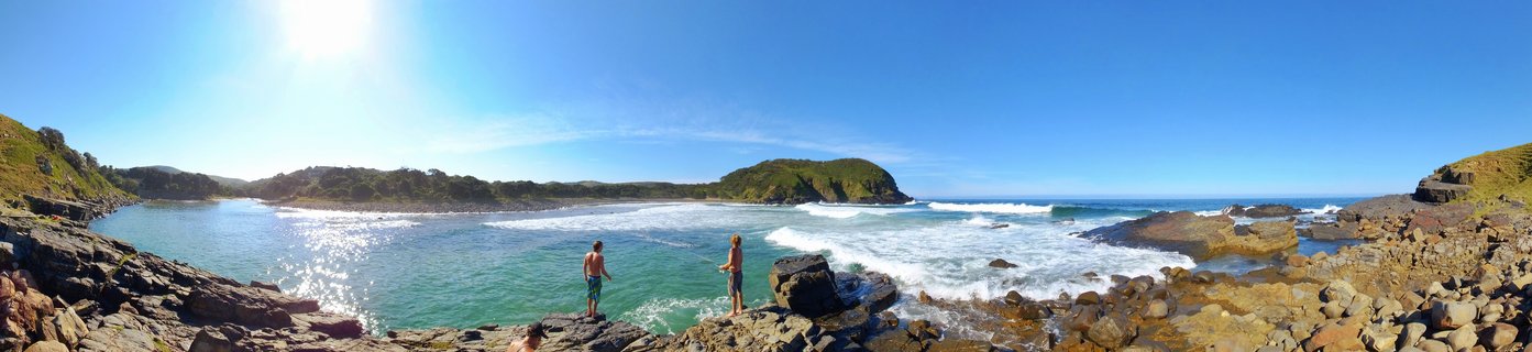 Panoramic view of two people standing on rocky shore overlooking turquoise bay with breaking waves, green hills and bright blue sky at Coffee Bay, South Africa