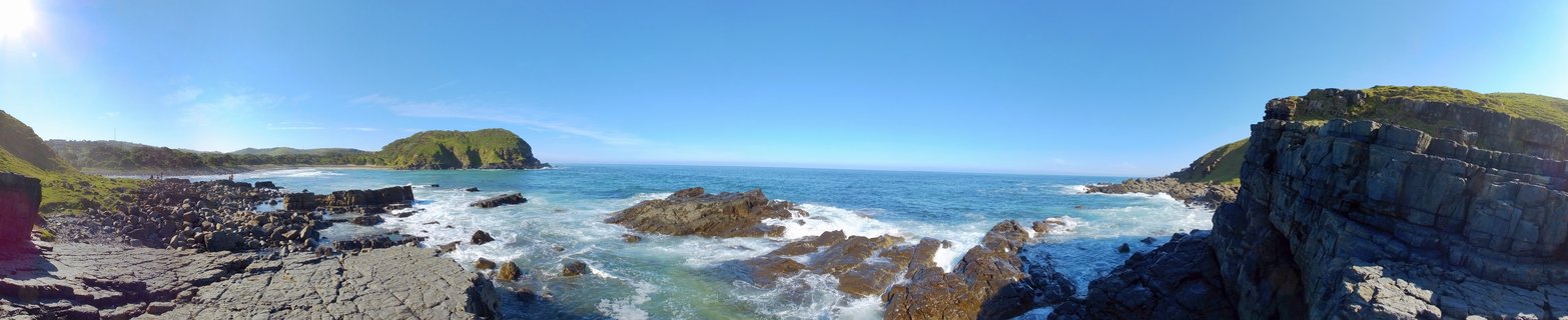 Panoramic view of rocky coastline with waves breaking over layered rock formations, green hills on both sides, blue ocean and clear sky at Coffee Bay, South Africa