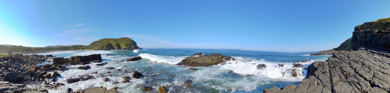 Panoramic view of rocky coastline with white waves crashing against dark grey rock formations, turquoise ocean water, green vegetated hills in the distance, and clear blue sky