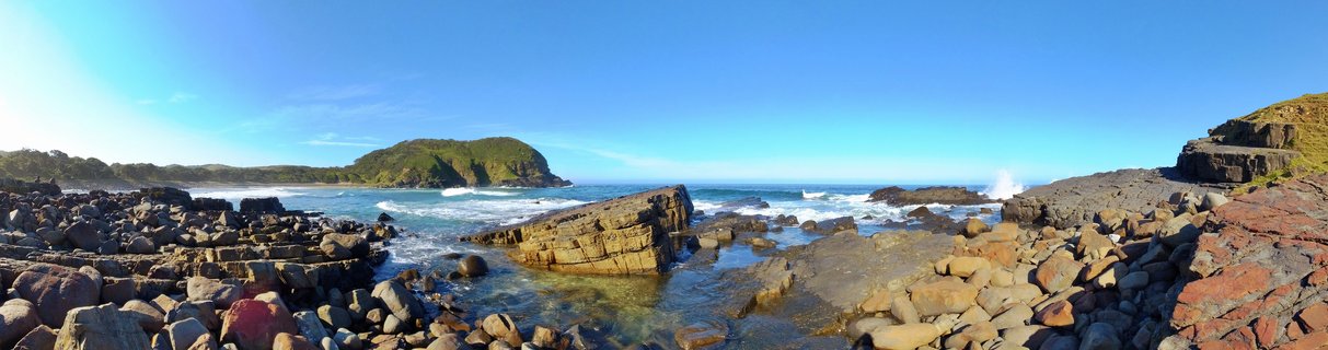 Panoramic coastal landscape showing rocky shoreline with layered sedimentary formations, ocean waves, green headland, and clear blue sky at Coffee Bay, South Africa