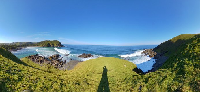 Panoramic coastal landscape showing green grassy hills overlooking blue ocean with white waves breaking on rocky shores under clear sky at Coffee Bay, South Africa
