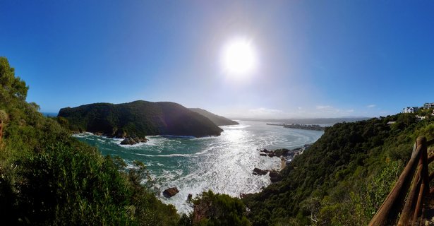 Panoramic coastal landscape showing the Knysna Heads headlands covered in green vegetation, with turquoise ocean water, white waves, rocky outcrops, and bright sun in clear blue sky