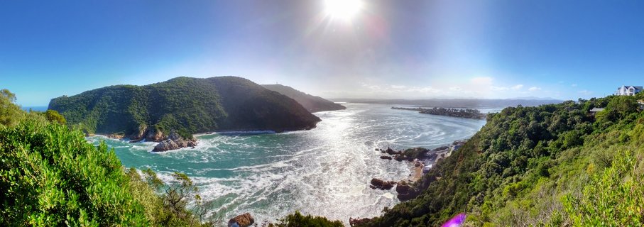 Panoramic coastal landscape showing a bay with turquoise water between two green forested hills, bright sun in blue sky, rocky shoreline with white waves, South Africa
