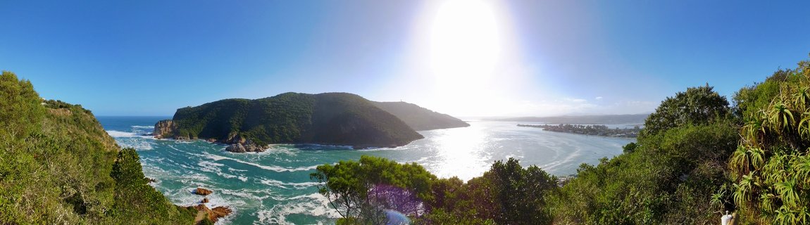Panoramic coastal landscape showing green headland jutting into turquoise ocean waters with bright sun overhead, lush vegetation in foreground, and distant town visible across calm lagoon waters