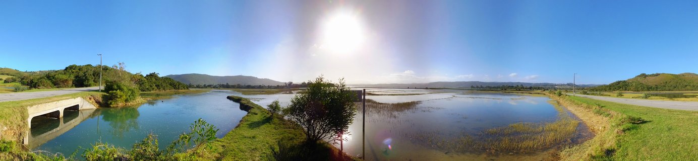 Wide panoramic landscape showing Knysna estuary wetlands with calm water reflecting blue sky, green grass and shrubs, concrete culvert bridge, distant mountains, and bright sun overhead