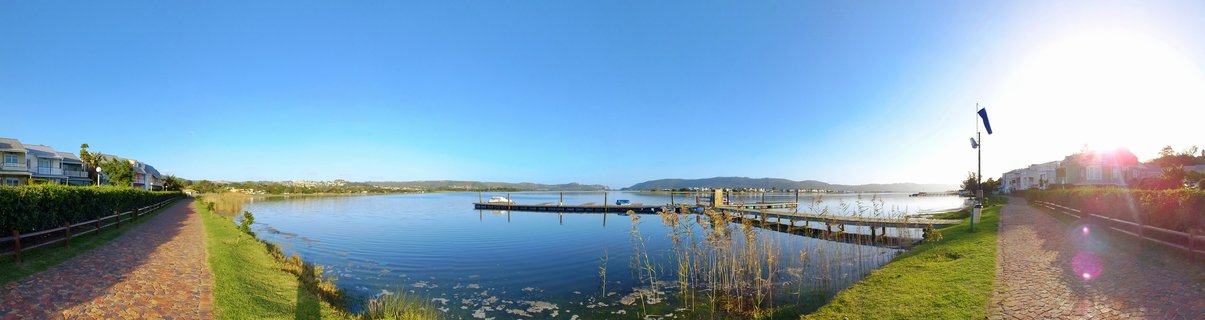 Panoramic waterfront scene showing a wooden pier extending into Knysna Lagoon with boats, residential buildings on both sides, paved walkways, and distant mountains under clear blue sky