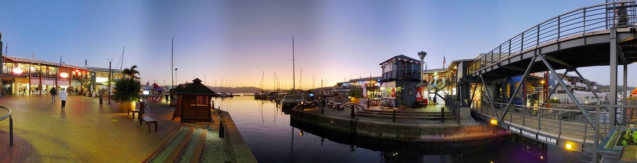Panoramic view of Knysna Waterfront marina at dusk showing calm harbor water with reflections, moored sailboats, illuminated two-story waterfront buildings with restaurants and shops, curved brick promenade with pedestrians walking, wooden gazebo, and modern steel pedestrian bridge on the right side under a twilight sky with orange and purple gradient