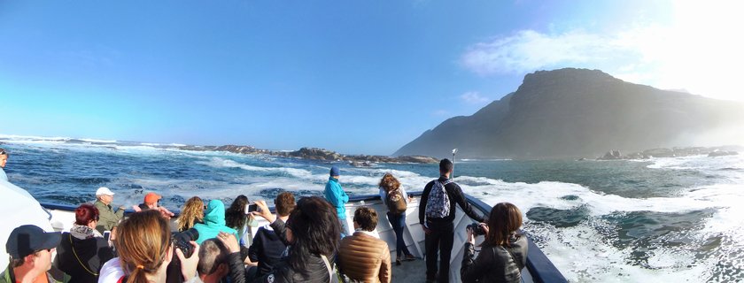 Group of tourists on a boat deck photographing Table Mountain and rocky coastline in Cape Town, South Africa under clear blue sky