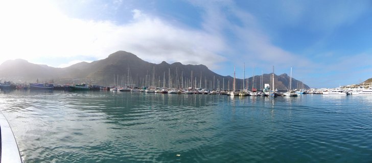 Panoramic view of a harbor filled with docked sailboats and yachts on calm blue-green water, with large mountains in the background under a blue sky with white clouds