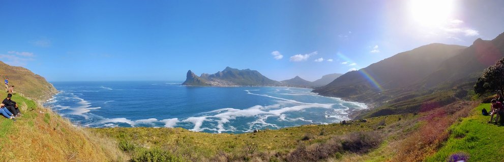 Panoramic coastal landscape showing Lion's Head mountain, Atlantic Ocean with breaking waves, and grassy hillsides with visitors at a viewpoint in Cape Town, South Africa