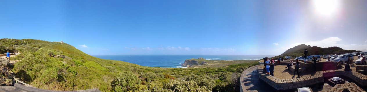 Panoramic view of Cape Point coastline with green hills, blue ocean, rocky cliffs, and tourists at a stone viewing platform under clear sky