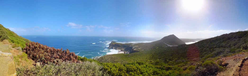 Wide panoramic coastal landscape showing green mountain slopes covered in vegetation descending to blue ocean with white waves, rocky headlands, and bright sun in clear sky over Cape Point, South Africa