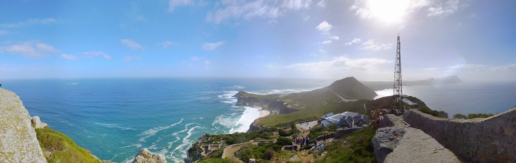 Wide panoramic view from Cape Point showing turquoise Atlantic Ocean, rocky coastline with white surf, green fynbos vegetation, stone viewing walls, visitor center buildings, tall communications tower, and bright sun in blue sky with scattered clouds