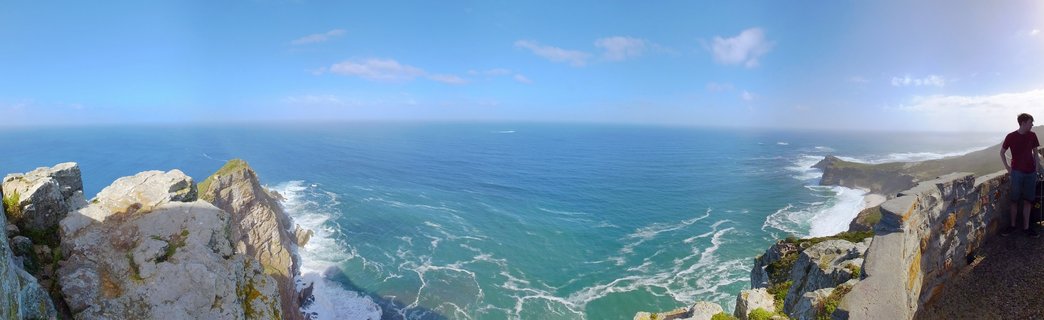 Panoramic coastal landscape showing dramatic rocky cliffs descending into turquoise Atlantic Ocean with white waves, blue sky with scattered clouds, and a person standing at a stone viewing wall on the right side