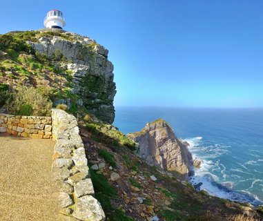 White lighthouse with red trim atop steep rocky cliff overlooking blue Atlantic Ocean, stone pathway and wall in foreground, Cape Point, South Africa