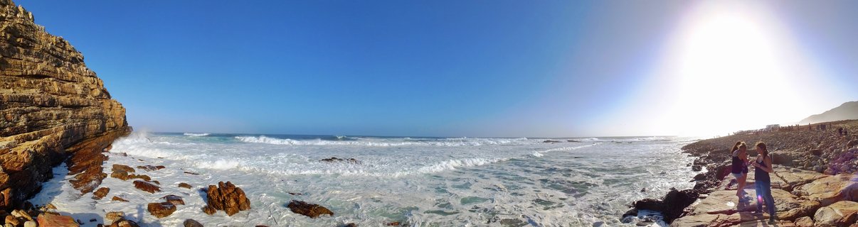 Coastal panorama showing layered rock cliffs, crashing ocean waves, and two people standing on rocky shore under bright sun and blue sky in Cape Town