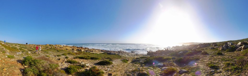 Wide coastal landscape showing rocky shoreline with green vegetation, ocean waves breaking on shore, bright sun in clear blue sky, and several people walking on coastal path