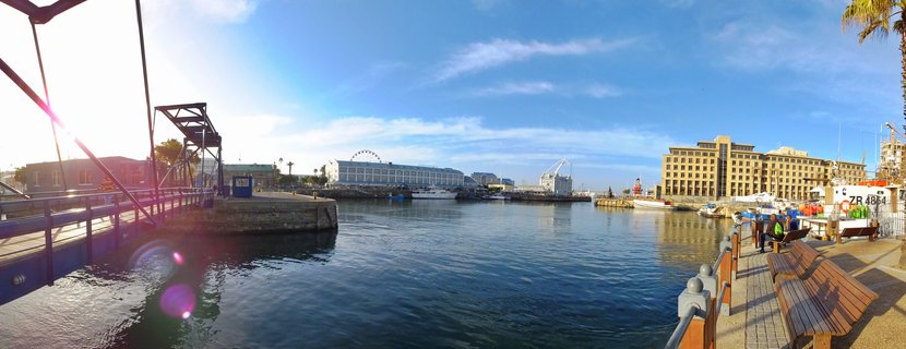 Panoramic view of V&A Waterfront harbor in Cape Town showing calm blue water, docked boats, waterfront buildings, Ferris wheel in distance, wooden pier structure, and benches along walkway under blue sky with afternoon sun