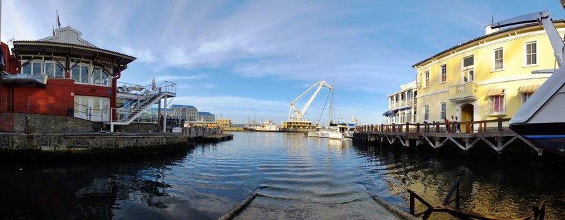 Harbor scene showing red and yellow historic buildings on either side of a marina with boats, water, and a crane visible in the distance under a blue sky with clouds in Cape Town, South Africa
