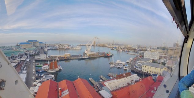 Elevated wide view of Cape Town's V&A Waterfront showing harbor with boats, cranes, red-roofed buildings, waterfront promenade, and blue sky with wispy clouds