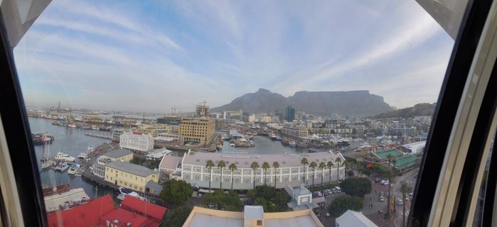 Panoramic view of Cape Town's V&A Waterfront showing harbor with boats, waterfront buildings with palm trees, and Table Mountain in the background under a partly cloudy sky
