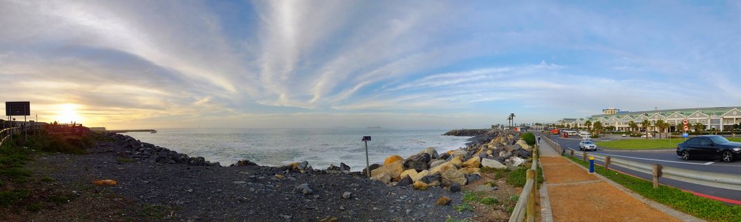 Coastal promenade in Cape Town, South Africa showing rocky shoreline, paved walkway, road with cars, and commercial buildings along the Atlantic Ocean during sunset with dramatic cloud formations