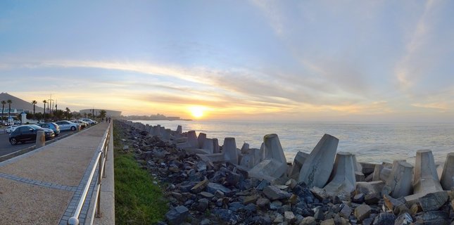 Sunset view over the Atlantic Ocean at Sea Point, Cape Town, showing concrete wave breakers along a rocky breakwater with a paved promenade, parked cars, and distant mountains under a golden sky