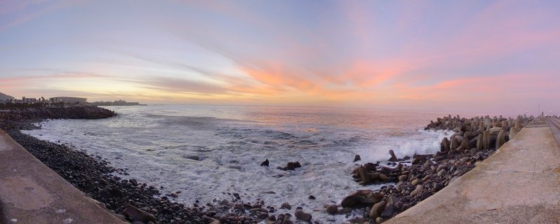 Panoramic view of ocean waves breaking against rock and concrete breakwater structures at sunset in Cape Town, South Africa, with colorful sky