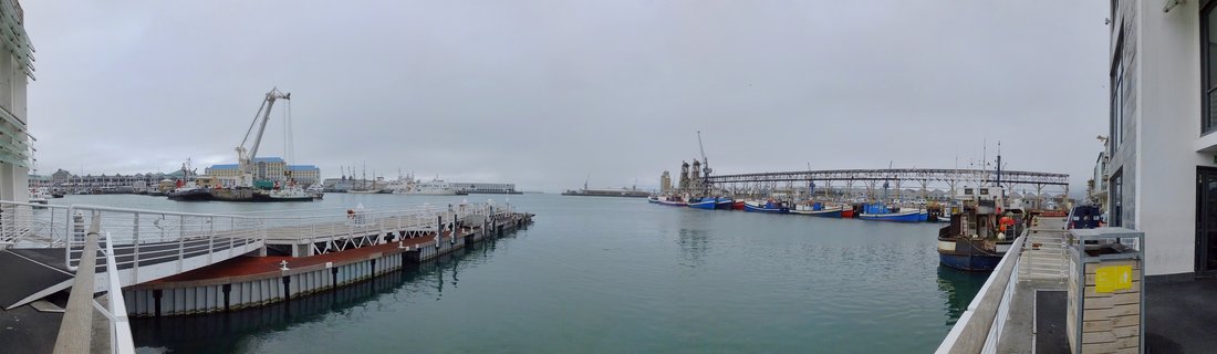 Harbor scene showing fishing boats docked at piers with industrial cranes and port infrastructure, calm water in center, buildings framing both sides under overcast sky