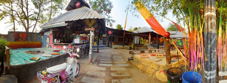Outdoor bar and restaurant in Thailand with a pink motorbike parked in front, colorful painted bamboo poles on the right, corrugated metal roof structures with murals, outdoor seating with cushions, and Buddhist prayer flags