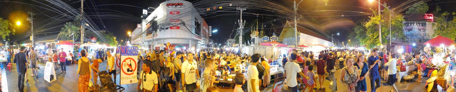 A crowded night market street in Chiang Mai, Thailand with numerous people browsing vendor stalls under illuminated canopies, with a Central department store building visible in the background