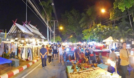 Night market street in Chiang Mai with illuminated vendor stalls selling clothing and handicrafts, people browsing and shopping, Thai-style architecture visible on the left, palm trees in background