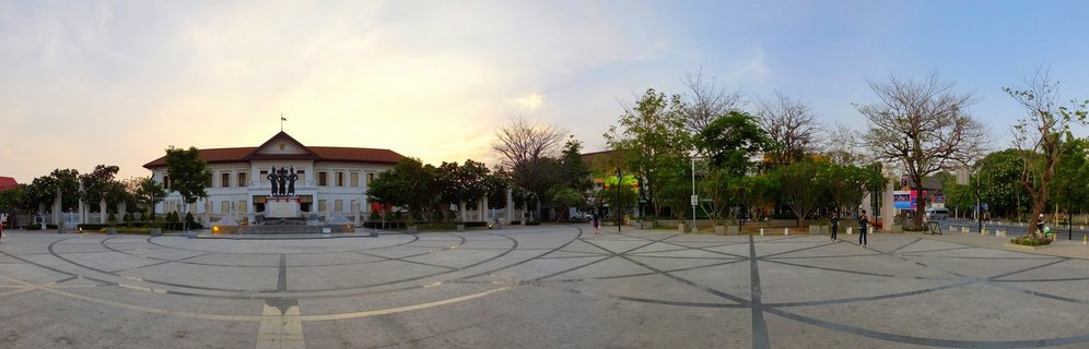 Wide plaza with circular pavement patterns featuring Three Kings Monument statue in front of white colonial building with red roof, trees and pedestrians at sunset in Chiang Mai Thailand