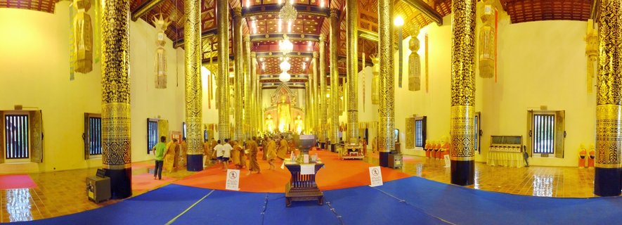 Wide interior view of a Thai Buddhist temple with multiple ornate golden pillars, monks in orange robes walking on red and blue carpets, and a golden Buddha statue visible at the far end of the hall