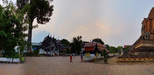Wide view of Wat Chedi Luang Buddhist temple courtyard in Chiang Mai with traditional Thai temple buildings, large brick stupa, monks in orange robes, and visitors walking on paved ground at dusk