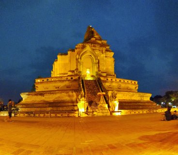 Large ancient Buddhist stupa illuminated with golden lights at dusk, with stone staircase flanked by serpent statues leading to an arched entrance, deep blue sky above