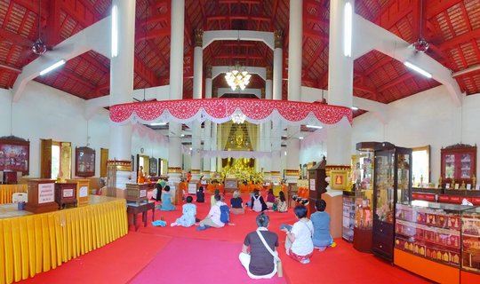 Interior view of a Buddhist temple prayer hall with red carpeted floor, white pillars, and red wooden ceiling. Visitors and monks in orange robes sit facing a golden Buddha statue at the far end of the hall.