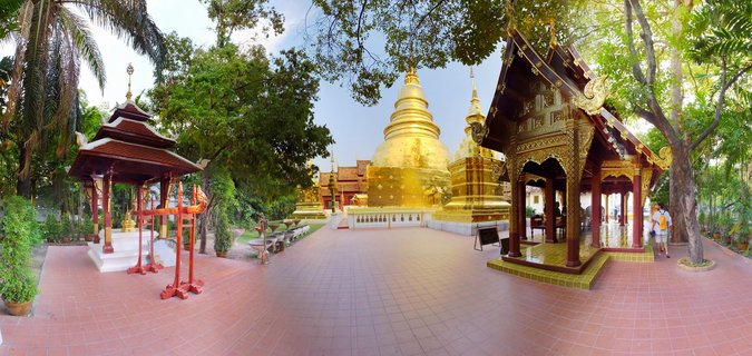 Panoramic photograph of a Thai Buddhist temple showing a large golden stupa in the center with two traditional wooden pavilions on either side, surrounded by palm trees and tropical vegetation on a tiled courtyard