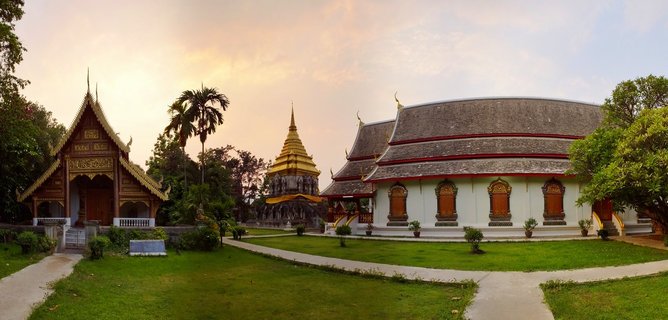Thai Buddhist temple complex with golden stupa between two traditional temple buildings with ornate tiered roofs, green lawn, palm trees, sunset sky