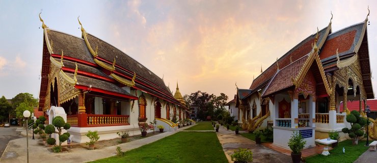 Two traditional Thai temple buildings with ornate golden roof decorations and multi-tiered roofs facing each other across a green lawn pathway under a sunset sky