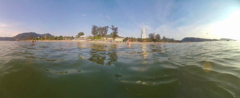 Split-level photograph showing ocean water in foreground with beach, swimmers, palm trees, hotels and mountains visible along the shoreline in Phuket, Thailand