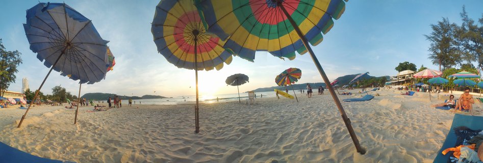 Wide beach panorama showing multiple colorful beach umbrellas on white sand with people relaxing, ocean waves, and sunset sky over Phuket Thailand