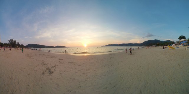 Wide beach scene at sunset in Phuket, Thailand showing sand, ocean, scattered people, distant mountains, and golden sky with sun low on the horizon over the water