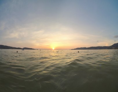 Sunset over ocean water with golden sky, silhouettes of people swimming, hills on horizon in Phuket Thailand