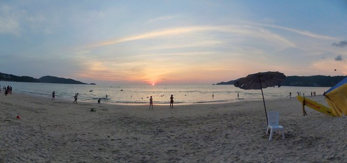 Wide beach scene at sunset in Phuket, Thailand, showing people on sand and in shallow water, with a beach umbrella, white chair, and yellow rescue board in the foreground, and hills visible across the bay