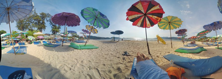 Wide beach scene showing multiple colorful striped umbrellas in blue purple green red and yellow with beach lounge chairs on white sand ocean in background blue sky above