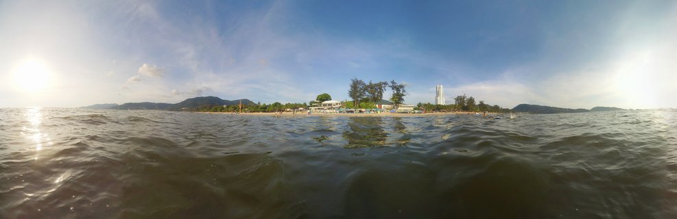 Panoramic seascape showing Patong Beach in Phuket with resort buildings, beach umbrellas, people on sand, coastal trees, mountains in background, and blue sky with clouds viewed from ocean water