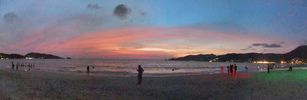 Wide beach scene at sunset showing people on sand with pink and orange sky, ocean waves, distant mountains and city lights along the coastline in Phuket Thailand