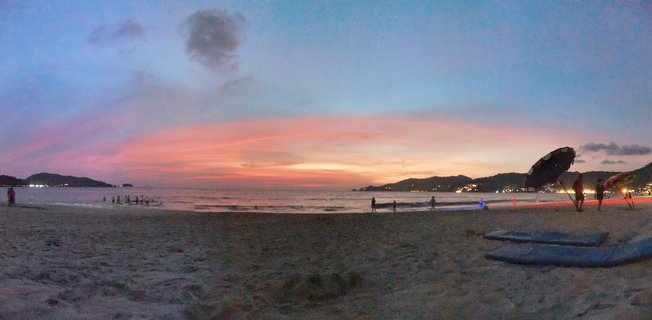 Wide beach scene at sunset showing sand foreground, ocean waves, silhouetted people, beach umbrellas, colorful pink and orange sky, and hills in the distance at Phuket Thailand