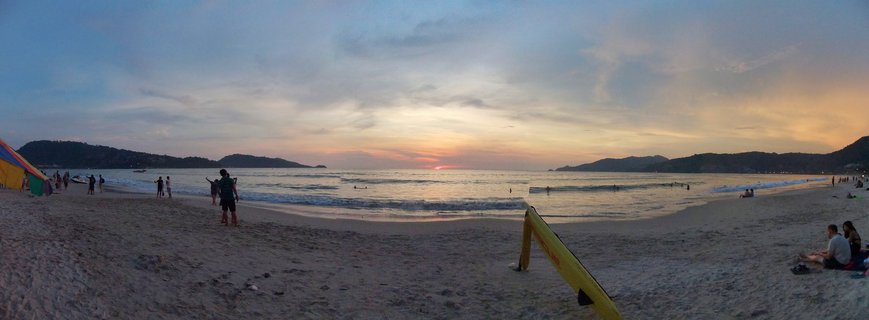 Panoramic view of a beach in Phuket at sunset with people on the sand, surfers in the water, hills in the background, and colorful sky with orange and blue tones